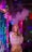 A person standing in a vibrant, colorful setting with neon lights and party decorations. The individual is holding a vaping device, exhaling a large cloud of vapor. The background is decorated with shiny, metallic streamers and balloons, creating a festive atmosphere.