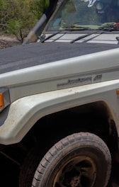 Close-up of the front section of a Toyota Land Cruiser 45 with visible dirt and scratches, parked in a natural outdoor setting. The headlight and fender are partially visible with a dusty tire in the foreground. Some vegetation is noticeable in the background.