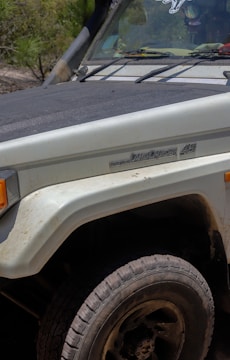 Close-up of the front section of a Toyota Land Cruiser 45 with visible dirt and scratches, parked in a natural outdoor setting. The headlight and fender are partially visible with a dusty tire in the foreground. Some vegetation is noticeable in the background.