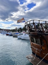 A wooden ship with an intricate railing is docked at a marina. The Croatian flag flies from the ship’s mast. Several modern boats are lined up along the waterfront, with palm trees and buildings in the background. The sky is partially cloudy with patches of blue visible.