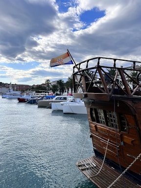 A wooden ship with an intricate railing is docked at a marina. The Croatian flag flies from the ship’s mast. Several modern boats are lined up along the waterfront, with palm trees and buildings in the background. The sky is partially cloudy with patches of blue visible.
