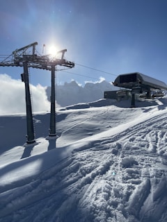 The image captures a snowy mountain landscape under clear blue skies. A ski lift with metal structures and cables is prominently featured, with the sun shining brightly behind it. The ground is covered in smooth, untouched snow, except for a few tracks, suggesting recent skiing or snowboarding activity.