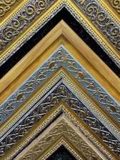 A collection of intricately designed, ornate picture frames arranged in a pattern. The frames exhibit detailed, swirling patterns with gold, silver, and black colors creating a striking visual contrast. The repetitive geometric layout emphasizes the craftsmanship and artistry.
