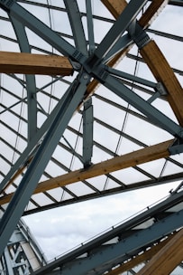 A complex architectural structure featuring intersecting metal beams and wooden accents. The framework is overlaid with large glass panels, allowing natural light to filter through.