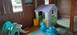 A children's play area with a small playhouse featuring a purple roof and white walls. There is a blue plastic rocking toy shaped like a horse in the foreground. A slide with a steering wheel is positioned to the left, and colorful interlocking foam mats cover the floor. The surroundings include wooden walls and an outdoor view through the window.