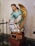 A statue of an angel with golden wings positioned on a pedestal. The figure is dressed in a turquoise robe with gold trim and has brown hair styled in braids. The angel appears to be holding a bag with inscriptions and is kneeling on a cloud-like structure.