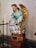 A statue of an angel with golden wings positioned on a pedestal. The figure is dressed in a turquoise robe with gold trim and has brown hair styled in braids. The angel appears to be holding a bag with inscriptions and is kneeling on a cloud-like structure.