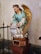 A statue of an angel with golden wings positioned on a pedestal. The figure is dressed in a turquoise robe with gold trim and has brown hair styled in braids. The angel appears to be holding a bag with inscriptions and is kneeling on a cloud-like structure.