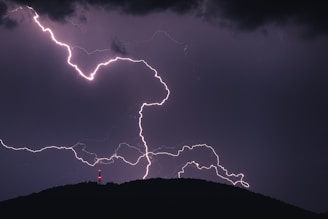 A powerful lightning strike illuminates the night sky, with jagged white streaks creating an intricate pattern. A dark silhouette of a hill is visible at the bottom, and a red light on a tower or structure adds a striking contrast to the scene.