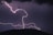 A powerful lightning strike illuminates the night sky, with jagged white streaks creating an intricate pattern. A dark silhouette of a hill is visible at the bottom, and a red light on a tower or structure adds a striking contrast to the scene.