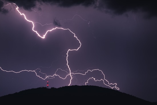 A powerful lightning strike illuminates the night sky, with jagged white streaks creating an intricate pattern. A dark silhouette of a hill is visible at the bottom, and a red light on a tower or structure adds a striking contrast to the scene.