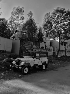 A vintage-style jeep is parked on the side of a dirt road, surrounded by trees and a building in the background. The scene is captured in grayscale, emphasizing the textures of the vehicle and the natural surroundings.
