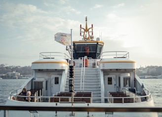 A ferry with a white and yellow design is on the water. There are steps leading up to an enclosed cabin. A flag is flying atop a pole, and life preservers are visible. Two people are seated on the lower deck, surrounded by empty benches. The background shows a city skyline with several buildings and trees under a partially cloudy sky.