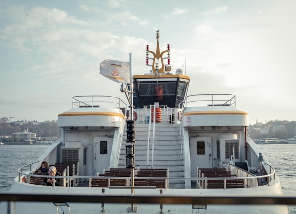 A ferry with a white and yellow design is on the water. There are steps leading up to an enclosed cabin. A flag is flying atop a pole, and life preservers are visible. Two people are seated on the lower deck, surrounded by empty benches. The background shows a city skyline with several buildings and trees under a partially cloudy sky.
