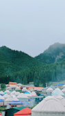 A collection of yurts with colorful roofs are nestled in a lush green forested valley, surrounded by misty mountains under an overcast sky.