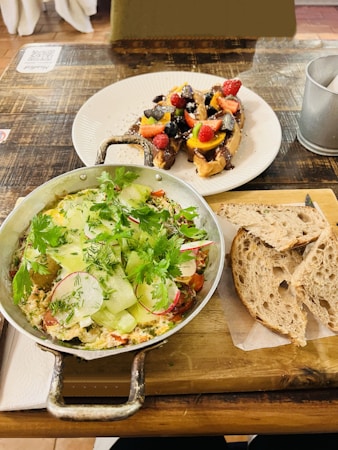 A table displaying a meal with two main dishes. The first dish is a colorful skillet filled with vegetables and herbs, possibly an omelet or frittata, topped with greens such as cilantro and thinly sliced cucumbers and radishes. Next to it, slices of rustic bread are placed on a wooden board. The second dish is a plate with toast topped with a variety of fresh fruits, including strawberries, raspberries, blueberries, and kiwi, possibly drizzled with chocolate or syrup.