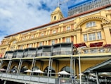 A large, historic building with classic architectural details such as arches and columns. The facade is painted in shades of yellow and dark red, featuring several arched windows. At the street level, there are outdoor seating areas with tables and umbrellas. Above, there are several balconies, and the upper parts of the building are under renovation with visible scaffolding. The sky is partly cloudy, providing a bright and airy backdrop.