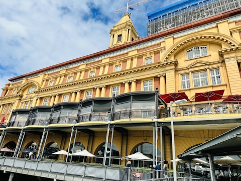 A large, historic building with classic architectural details such as arches and columns. The facade is painted in shades of yellow and dark red, featuring several arched windows. At the street level, there are outdoor seating areas with tables and umbrellas. Above, there are several balconies, and the upper parts of the building are under renovation with visible scaffolding. The sky is partly cloudy, providing a bright and airy backdrop.