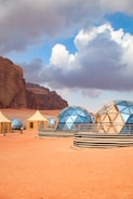 A desert landscape features several geodesic domes with reflective surfaces, situated on an arid, reddish-orange ground. Nearby, there are smaller, tent-like structures. Rocky cliffs rise majestically in the background under a dramatic sky filled with large, fluffy clouds.