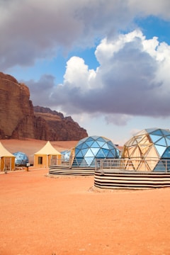 A desert landscape features several geodesic domes with reflective surfaces, situated on an arid, reddish-orange ground. Nearby, there are smaller, tent-like structures. Rocky cliffs rise majestically in the background under a dramatic sky filled with large, fluffy clouds.