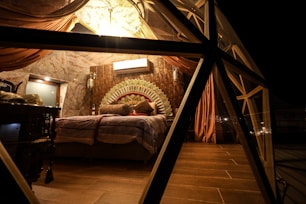 A cozy, dimly lit bedroom with a geometric glass structure. The room features a plush bed adorned with multiple cushions, a decorative headboard with an intricate pattern, and draped curtains hanging from the ceiling. The lighting is warm, creating a comfortable and inviting atmosphere.