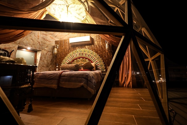 A cozy, dimly lit bedroom with a geometric glass structure. The room features a plush bed adorned with multiple cushions, a decorative headboard with an intricate pattern, and draped curtains hanging from the ceiling. The lighting is warm, creating a comfortable and inviting atmosphere.