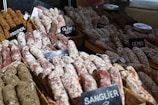 Various types of sausages are arranged in wooden trays, each with labels indicating flavors such as Provencal, Olives, and Cèpes. The sausages have a rustic appearance with a mix of dark and white coatings.