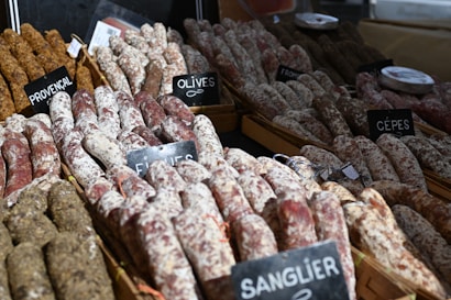 Various types of sausages are arranged in wooden trays, each with labels indicating flavors such as Provencal, Olives, and Cèpes. The sausages have a rustic appearance with a mix of dark and white coatings.