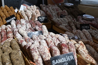 Various types of sausages are arranged in wooden trays, each with labels indicating flavors such as Provencal, Olives, and Cèpes. The sausages have a rustic appearance with a mix of dark and white coatings.
