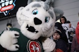 A large, plush mascot with a lion or tiger-like appearance is interacting with children and spectators. It has white fur with gray patches and large blue eyes. The background includes fans in winter clothing, some holding merchandise bags.