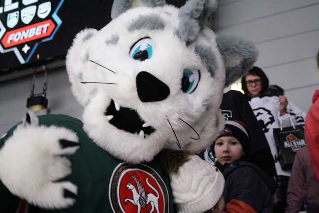 A large, plush mascot with a lion or tiger-like appearance is interacting with children and spectators. It has white fur with gray patches and large blue eyes. The background includes fans in winter clothing, some holding merchandise bags.