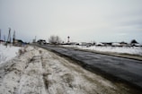 A rural road covered in patches of snow is seen winding through an open area with scattered houses in the distance. The sky is overcast, contributing to a subdued atmosphere. Utilities such as telephone poles line the road, and traffic signs are visible near the houses.