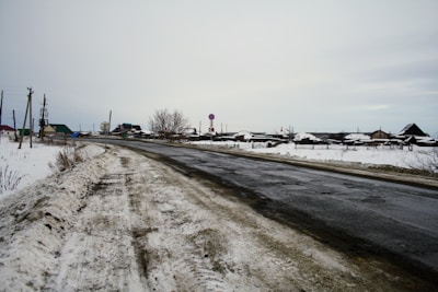 A rural road covered in patches of snow is seen winding through an open area with scattered houses in the distance. The sky is overcast, contributing to a subdued atmosphere. Utilities such as telephone poles line the road, and traffic signs are visible near the houses.
