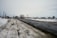 A rural road covered in patches of snow is seen winding through an open area with scattered houses in the distance. The sky is overcast, contributing to a subdued atmosphere. Utilities such as telephone poles line the road, and traffic signs are visible near the houses.