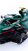 A green Formula 1 racing car with various sponsor logos including Aston Martin, Bombardier, JCB, Oakley, BWT, and SentinelOne. The car features intricate aerodynamic components and a sleek design. It has a single cockpit with a visible steering wheel and control mechanisms.
