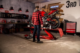 A person in a red and black checkered jacket is working on a motorcycle that is elevated on a red hydraulic lift in a garage. The garage is well-equipped with a tool rack on the wall, containing various tools and equipment organized neatly. There is a red tool chest on wheels to the right, and a sign on the wall says 'UNITED WE RIDE' along with a large '3rd'. Black and red pendant lights hang from the ceiling, adding to the workshop ambiance.
