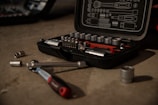A tool kit is open on a concrete surface, displaying various socket wrenches and attachments neatly arranged in a molded case. Several sockets and a ratchet handle are scattered around the case.