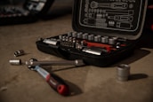 A tool kit is open on a concrete surface, displaying various socket wrenches and attachments neatly arranged in a molded case. Several sockets and a ratchet handle are scattered around the case.