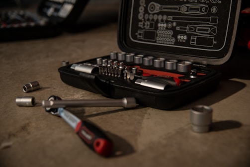 A tool kit is open on a concrete surface, displaying various socket wrenches and attachments neatly arranged in a molded case. Several sockets and a ratchet handle are scattered around the case.