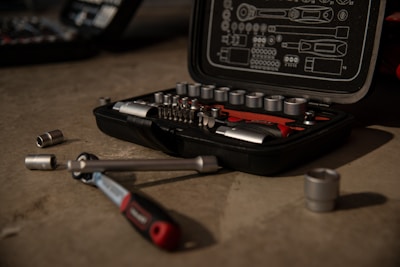A tool kit is open on a concrete surface, displaying various socket wrenches and attachments neatly arranged in a molded case. Several sockets and a ratchet handle are scattered around the case.