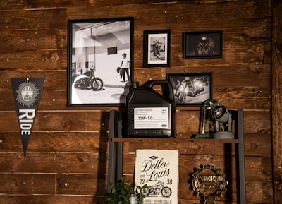 A wooden wall features a collection of framed black-and-white photographs depicting motorcycles and riders. A triangular flag with the word 'RIDE' hangs on the left side. A shelf holds a large container of motor oil alongside a trophy and a vintage-style sign.