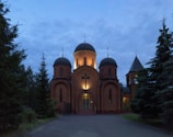 A church with illuminated domes stands amid tall evergreen trees under a dusky sky. Warm lights highlight the cross and architectural details of the structure, creating a serene and peaceful atmosphere.