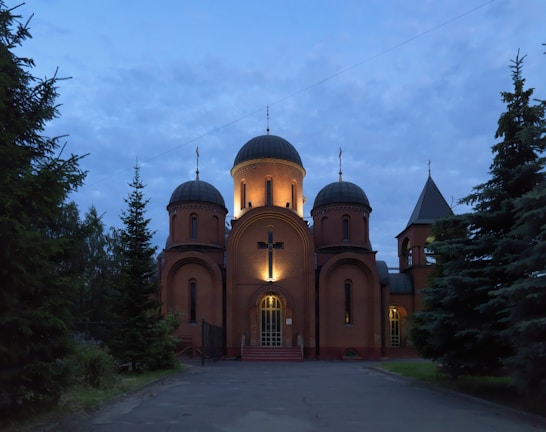 A church with illuminated domes stands amid tall evergreen trees under a dusky sky. Warm lights highlight the cross and architectural details of the structure, creating a serene and peaceful atmosphere.