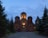 A church with illuminated domes stands amid tall evergreen trees under a dusky sky. Warm lights highlight the cross and architectural details of the structure, creating a serene and peaceful atmosphere.