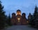 A church with illuminated domes stands amid tall evergreen trees under a dusky sky. Warm lights highlight the cross and architectural details of the structure, creating a serene and peaceful atmosphere.
