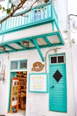 A charming storefront with a teal balcony and door, displaying a sign advertising a shop called 'Le Chocolat' and 'Matsouka', known for dry nuts, Mykonos delights, and Greek baklava. The interior is visible, showing shelves filled with various products. Leafy branches partially frame the scene.