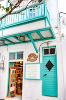 A charming storefront with a teal balcony and door, displaying a sign advertising a shop called 'Le Chocolat' and 'Matsouka', known for dry nuts, Mykonos delights, and Greek baklava. The interior is visible, showing shelves filled with various products. Leafy branches partially frame the scene.