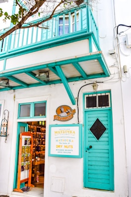 A charming storefront with a teal balcony and door, displaying a sign advertising a shop called 'Le Chocolat' and 'Matsouka', known for dry nuts, Mykonos delights, and Greek baklava. The interior is visible, showing shelves filled with various products. Leafy branches partially frame the scene.
