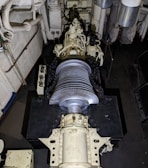 A large mechanical turbine with numerous metal blades is centrally positioned in a confined, industrial setting. Surrounding pipes, valves, and machinery suggest a technical or engineering environment, possibly within a power plant or ship engine room. The turbine features intricate details and components for mechanical operations.