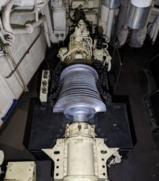 A large mechanical turbine with numerous metal blades is centrally positioned in a confined, industrial setting. Surrounding pipes, valves, and machinery suggest a technical or engineering environment, possibly within a power plant or ship engine room. The turbine features intricate details and components for mechanical operations.